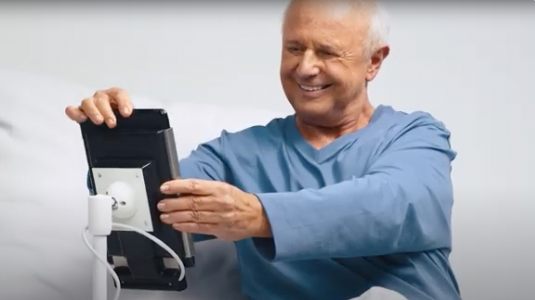 ConnectedCare.Komforthalter iPad – Älterer Patient im Krankenhausbett bedient lächelnd iPad an Nachttisch-Halterung