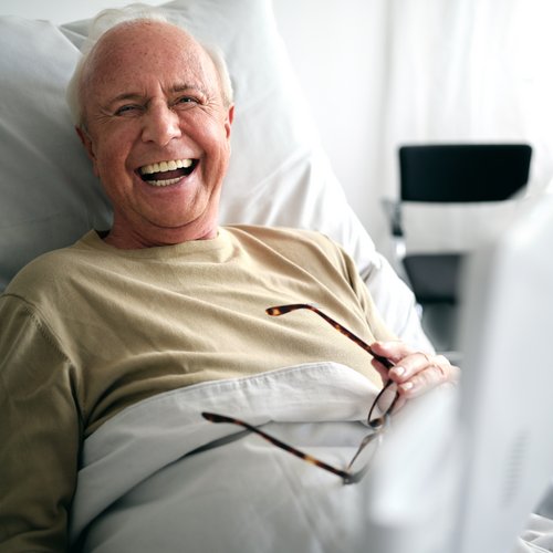 Älterer Patient im Krankenhausbett lacht herzhaft, BEWATEC Tablet unscharf im Vordergrund