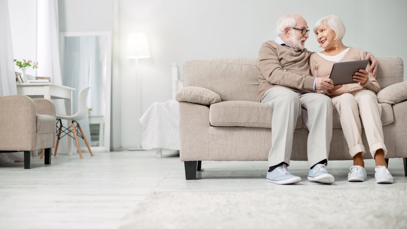 BEWATEC Third Party Partner | GOLDENSUMMER.TV, Senioren-Ehepaar sitzt Arm in Arm auf Sofa im Wohnzimmer mit Tablet