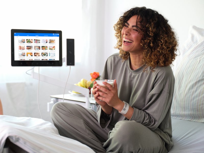 BEWATEC Patientenkommunikation | Patientin sitzt lachend im Bett mit einem Kaffee in der Hand