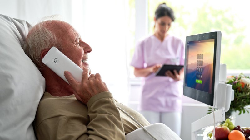 BEWATEC Patientenkommunikation | Patient liegt im Bett und telefoniert mit BEWATEC Phone, Pflegerin im Hintergrund