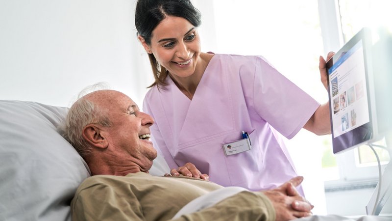 Patientenkommunikation: Junge Pflegerin und älterer Patient schauen fröhlich auf Bedside Terminal