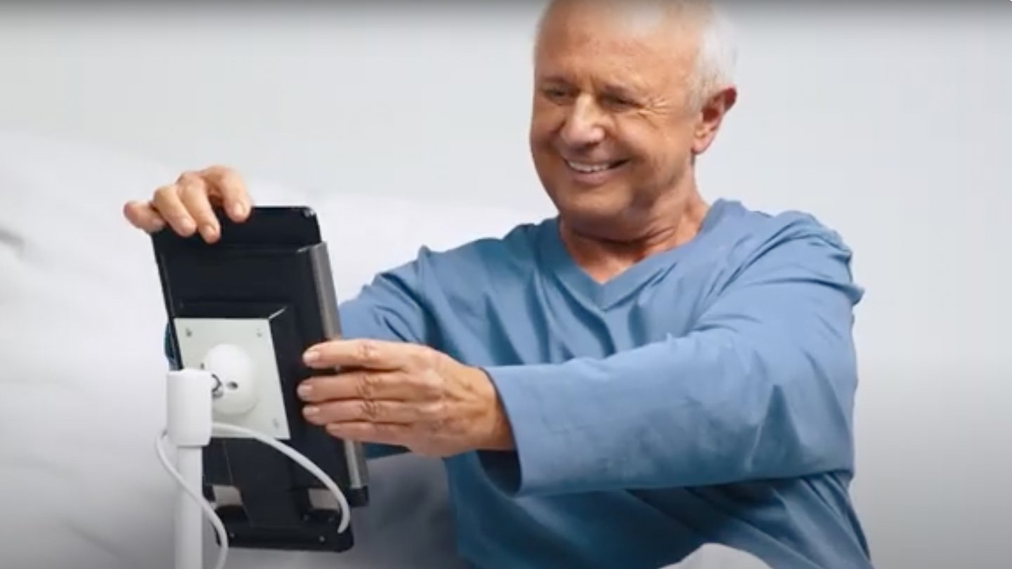 BEWATEC.Komforthalter iPad – Älterer Patient im Krankenhausbett bedient lächelnd iPad an Nachttisch-Halterung