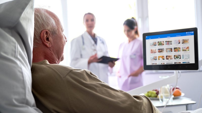 Älterer Patient sitzt im Krankenhausbett und schaut auf Bedside Terminal, Ärztin und Pflegerin im Hintergrund