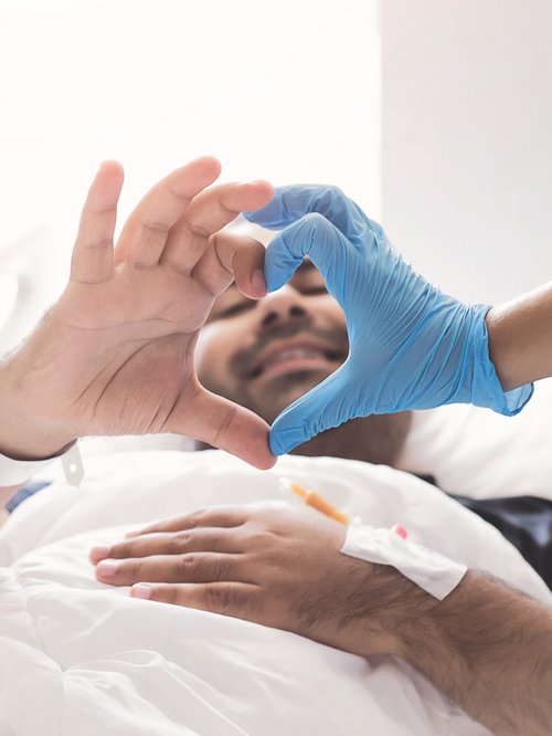 Patient liegt im Krankenhausbett und formt mit seiner rechten Hand und der Hand eines Klinikmitarbeitenden ein Herz.