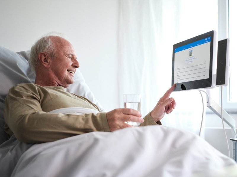 Patient Engagement | Älterer Patient liegt im Krankenhausbett und bedient Bedside Terminal