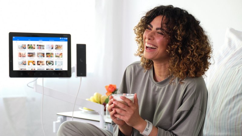 Patientenkommunikation: Patientin sitzt lachend im Bett mit einem Kaffee in der Hand