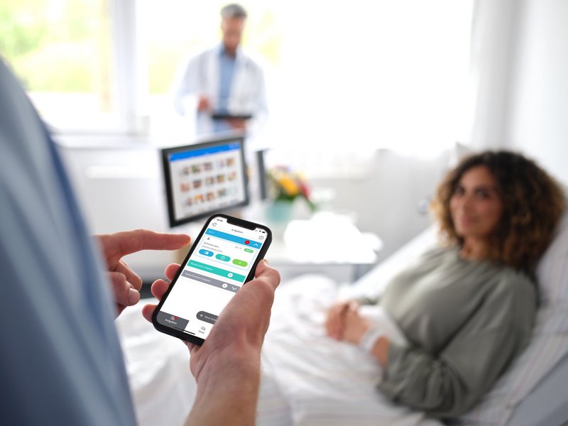 BEWATEC Patientenkommunikation | Patientin liegt im Bett und schaut zu Pfleger, der auf sein mit Smartphone mit BEWATEC.ConnectedCare schaut, Arzt mit Tablet im Hintergrund