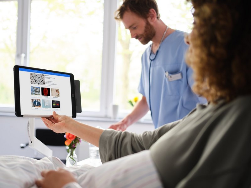 BEWATEC Krankenhaus digitalisieren | Patientin liegt im Bett und bedient iPad am, Pfleger im Hintergrund