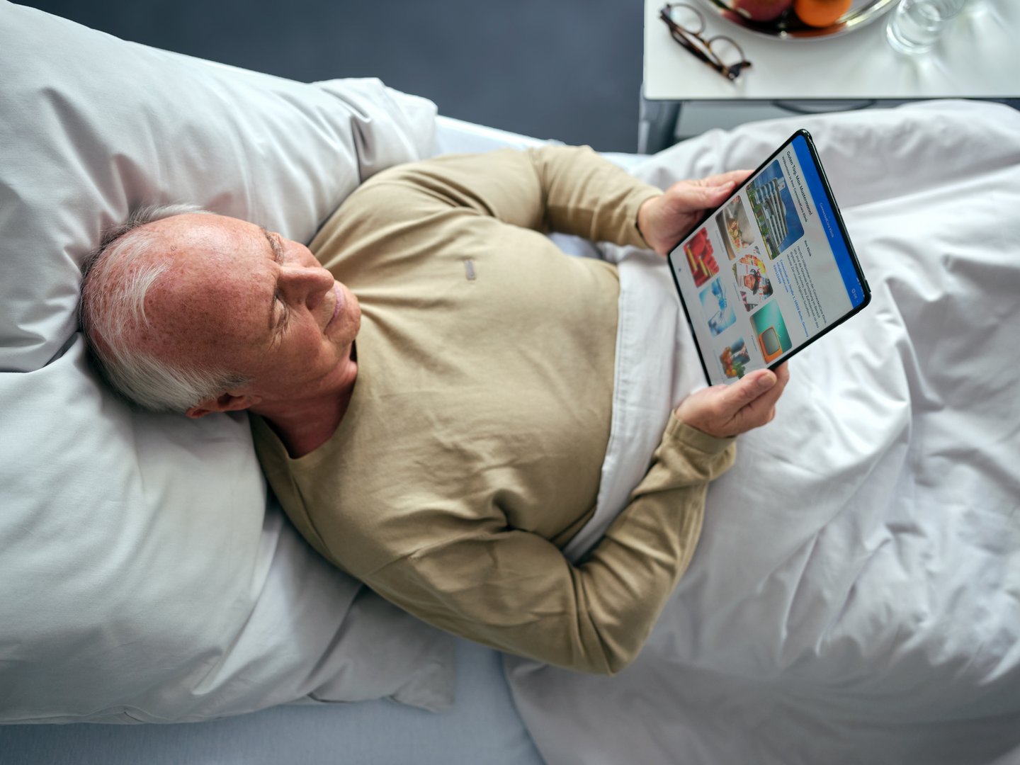 Patientenentertainment: Alterer Patient liegt im Krankenhausbett und liest auf dem iPad