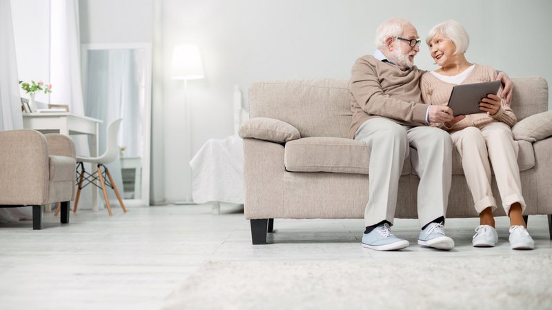 BEWATEC Third Party Partner | GOLDENSUMMER.TV, Senioren-Ehepaar sitzt Arm in Arm auf Sofa im Wohnzimmer mit Tablet